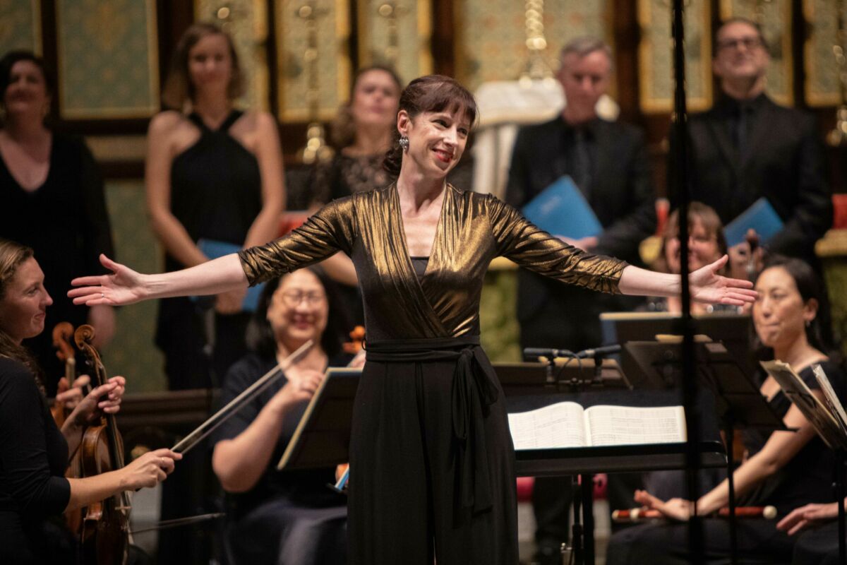 Madeleine Easton, Artistic Director of Bach Akademie Australia, standing in front of the orchestra with her arms outstretched in recognition of audience applause. She is wearing a sparkly gold top and a black dress.