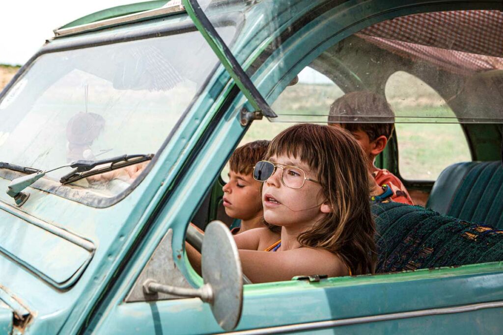 A little girl wears sunglasses with one of the lenses missing. She drives an old blue car with two young boys in it.
