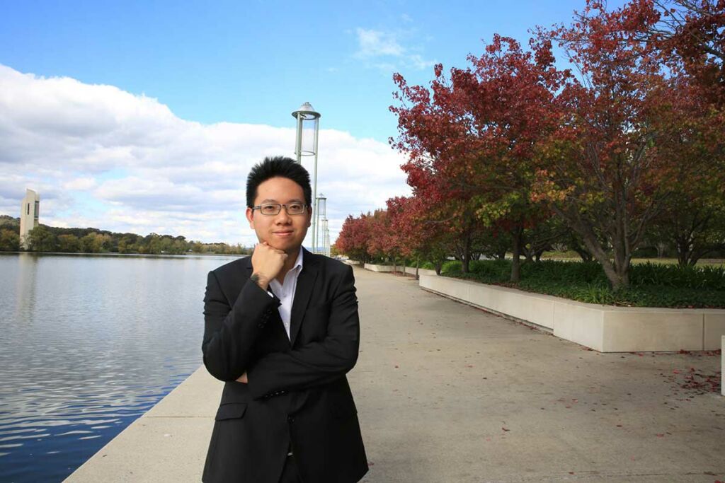 Alexander Yau stands on a walkway by a river with his fist under his chin.