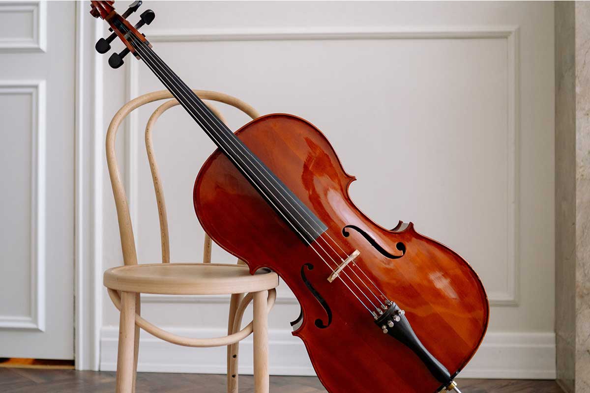 A cello leans against a white chair in front of a clean white wall.