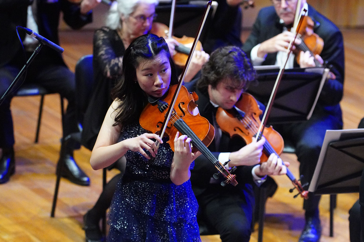 Hayley Lau in a blue dress, standing and performing on violin with a small section of the orchestra in the background.