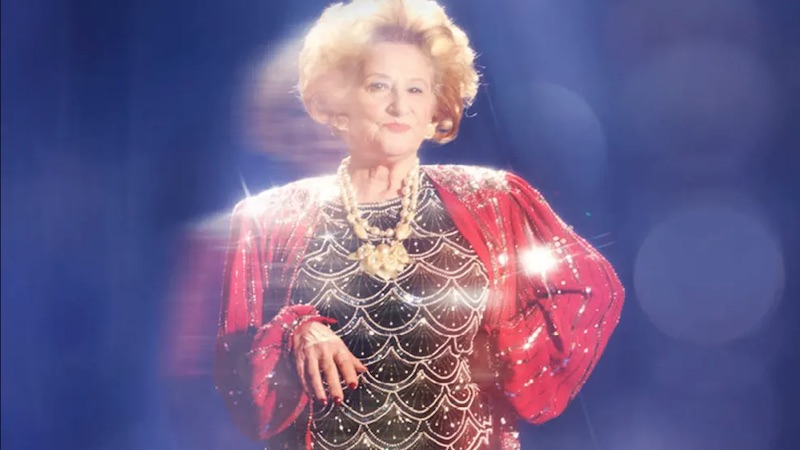 An older woman with big hair. Her shiny black shirt and red jacket catches the light.