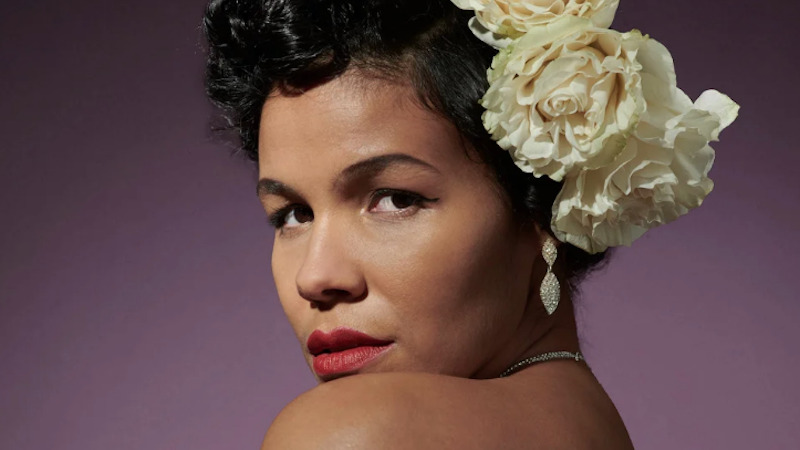 A woman dressed like Billie Holiday with white roses in her hair looks over her shoulder into the camera.