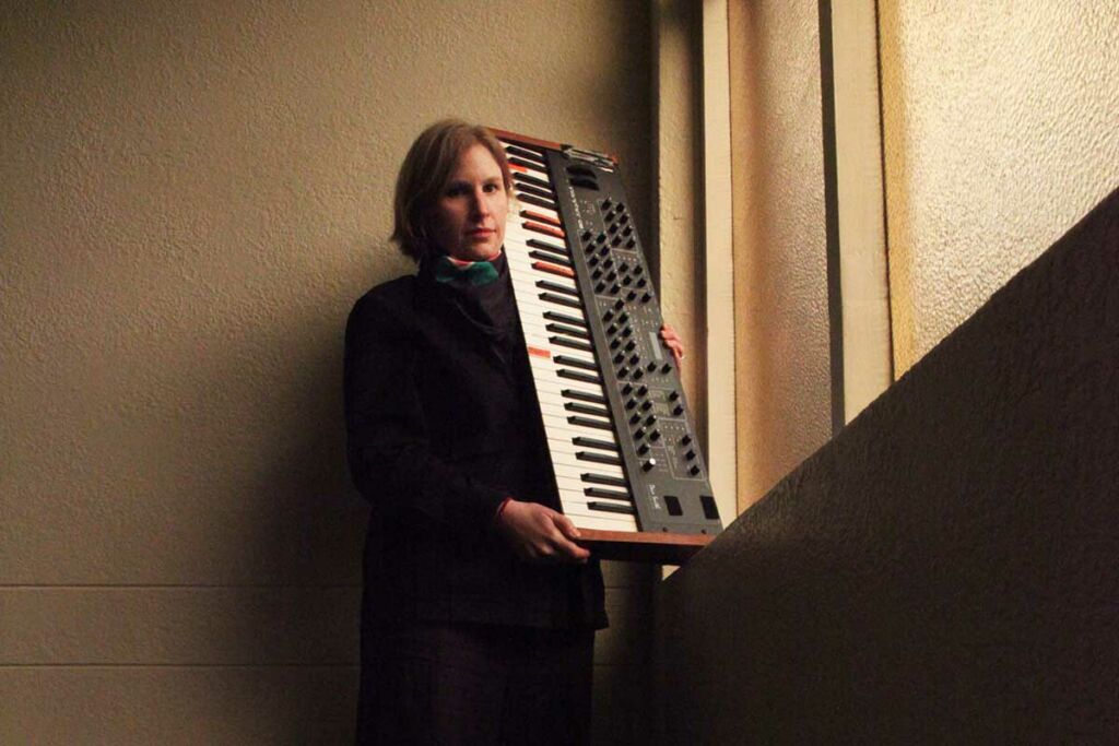 Heather Shannon holds a synthesiser upright over her shoulder. She stands next to a window.
