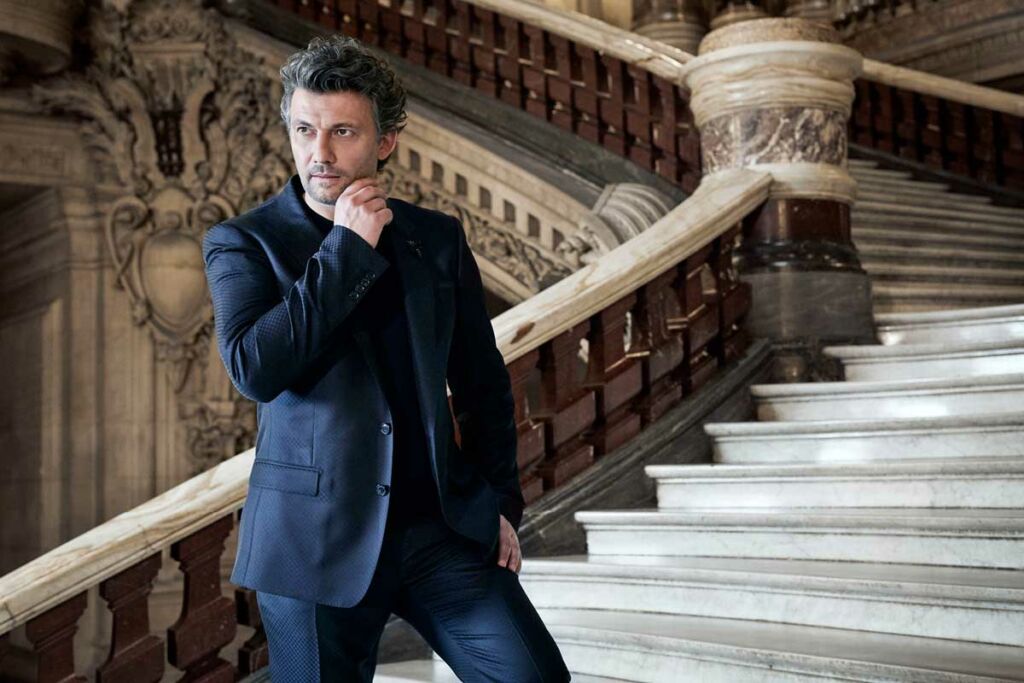 Jonas Kaufmann stands on a grand staircase in a suit, his hand on his chin.