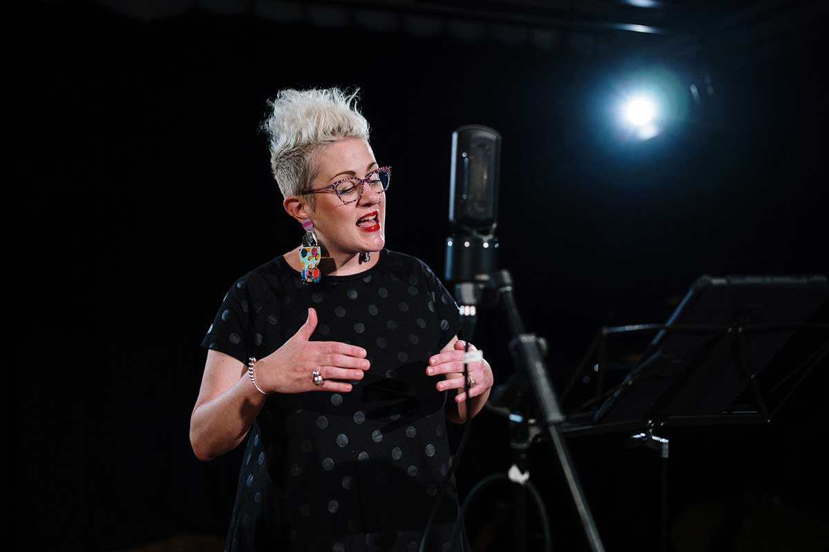 Katie Noonan sings into a microphone in a dark recording studio.