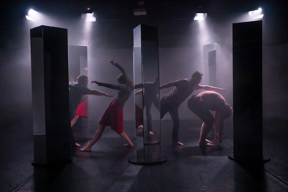 Two pairs of dancers interact with each other on a set of mirrors. One person pushes another back without touching, another curls up as a person extends their hand above their back.