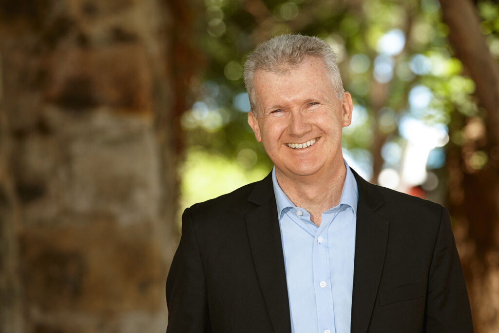 Tony Burke smiles, backdropped by trees and a sandstone brick wall.
