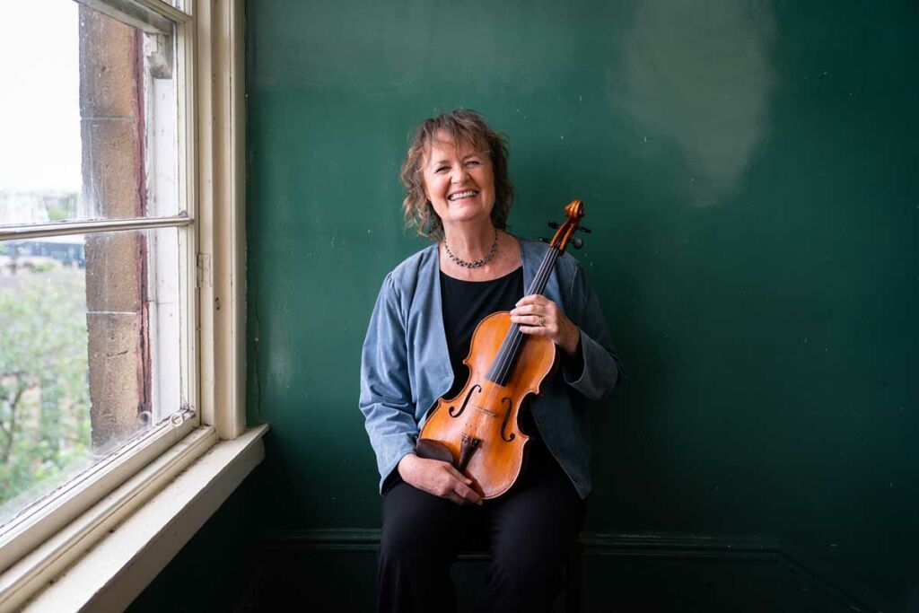 Sally Beamish holds her viola against a dark green wall, next to a window.