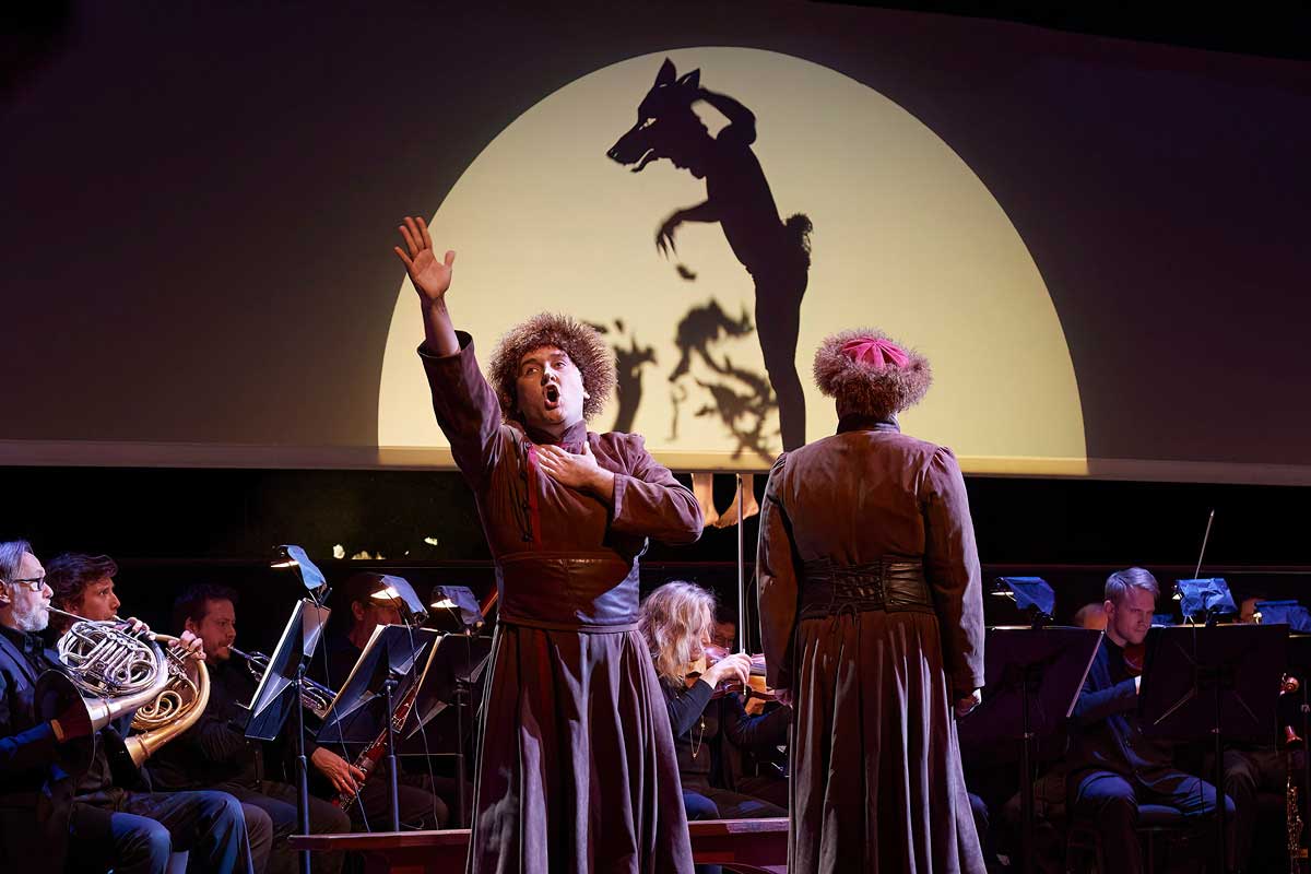 A man sings in front of an orchestra in, with the silhouette of a man with a wolf's head displayed in the background.