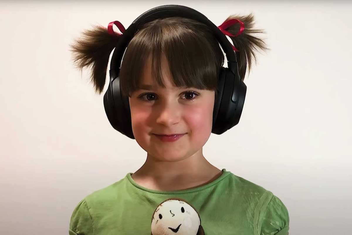 A little girl with short pigtails and headphones on holds her doll and smiles.
