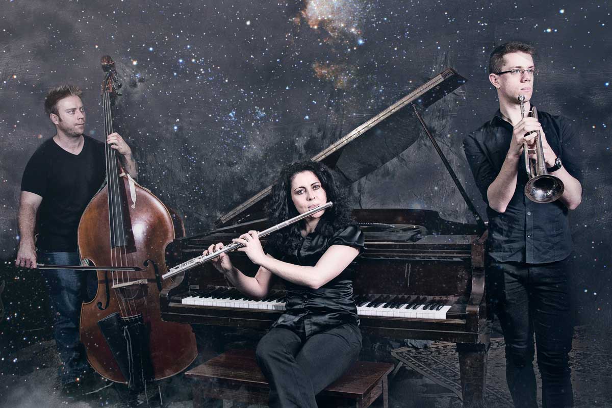Elsen Price holds his double bass, Keyna Wilkins plays flute while sitting in front of a piano, Will Gilbert holds his trumpet. The backdrop is starry space.