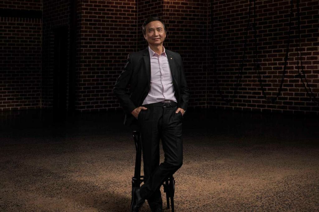 Li Cunxin sits on a stool under a spotlight, with dark brick walls behind him.