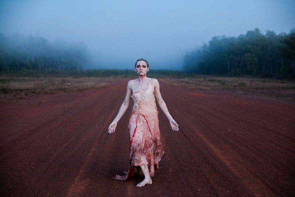 A dancer stands in the middle of a red dirt road in the fog, covered in white body paint and in a scaly pale dress.