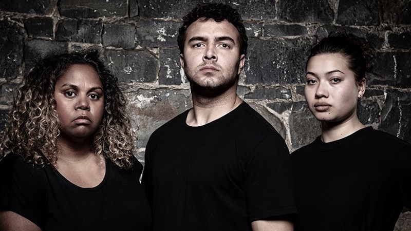 Three people in black shirts against a brick wall look at the camera with a hint of sadness.