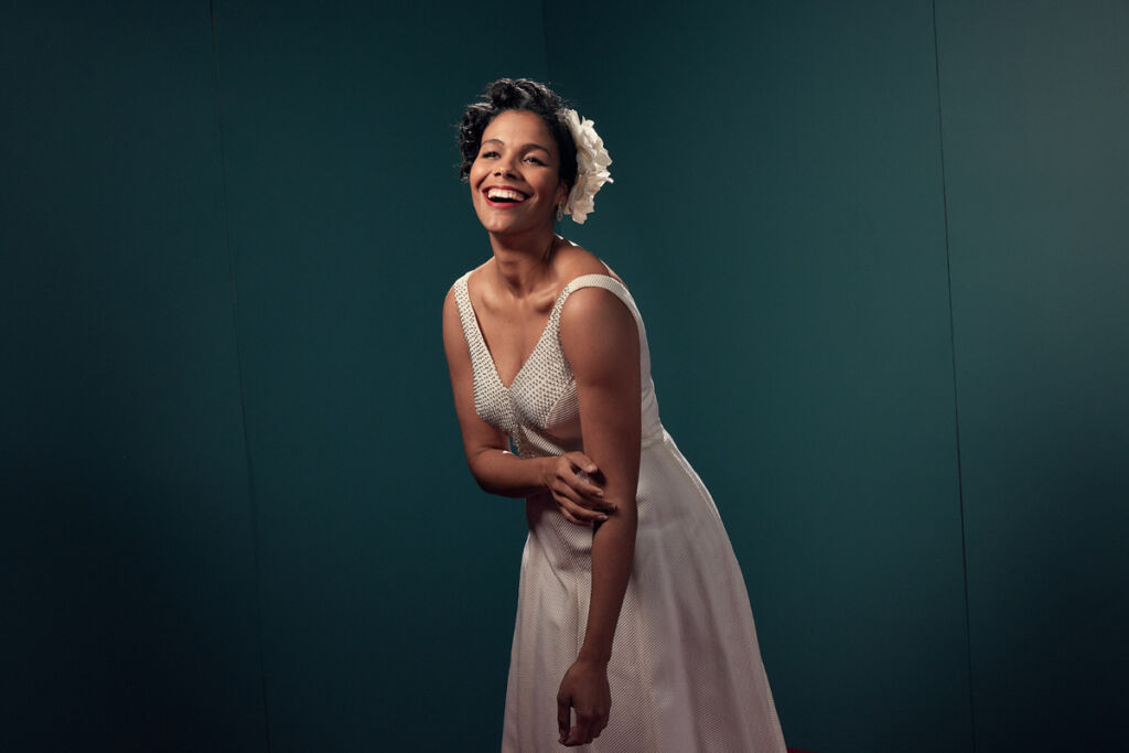 Zahra Newman, dressed as Billie Holiday in a 40s white dress and flowers in her hearm smiles.
