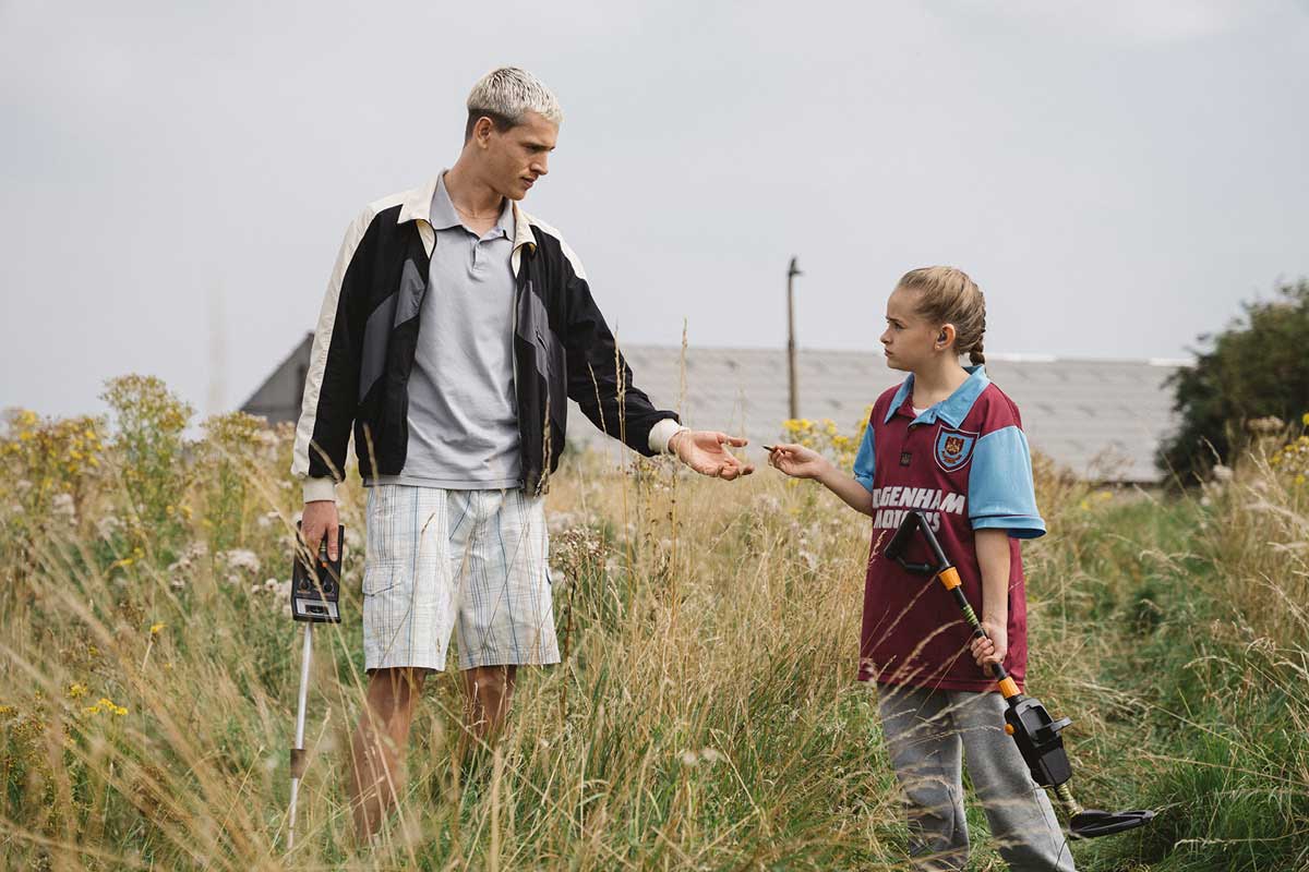 A young girl in a soccer uniform stands with a young man in a field. They both hold metal detectors, and the young girl is passing a small object to him