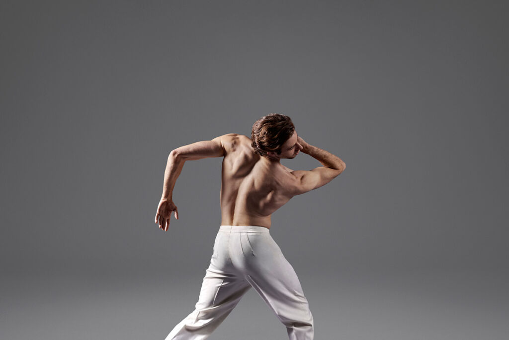 A dancer has his back turned to the camera, in motion. He is wearing white pants and no shirt.
