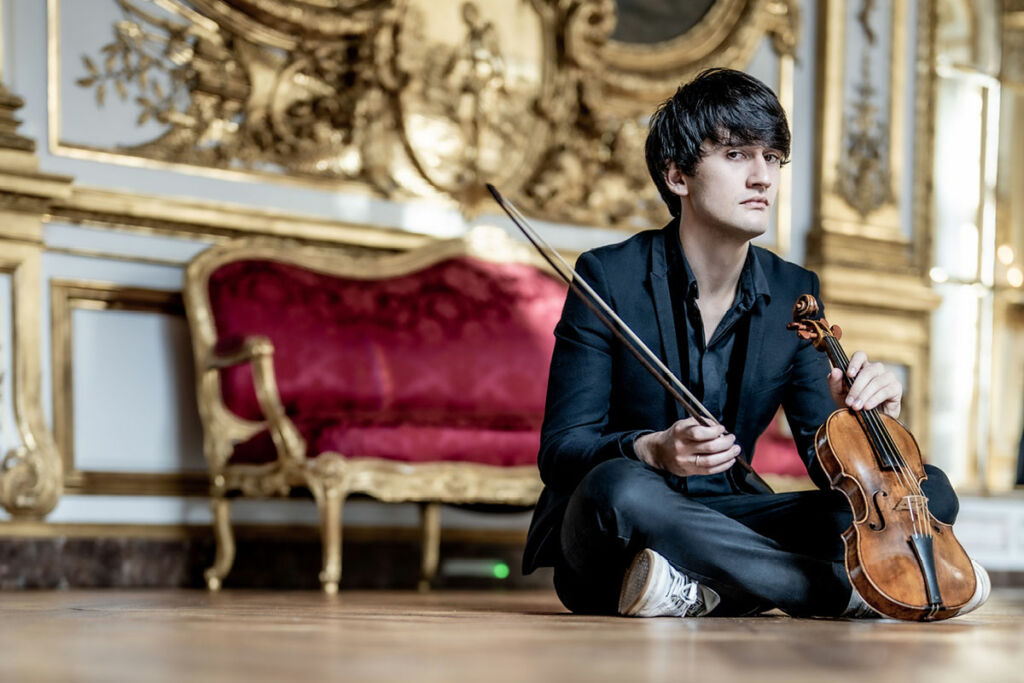 Théotime Langlois de Swarte sits on the floor in an ornately decorated room, holding his violin.