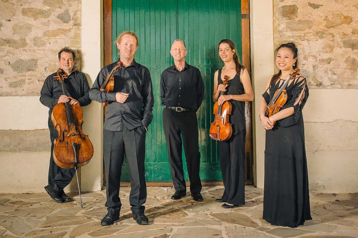 A string quintet stand in front of a sandstone wall and green door, holding their instruments – except the pianist, of course.