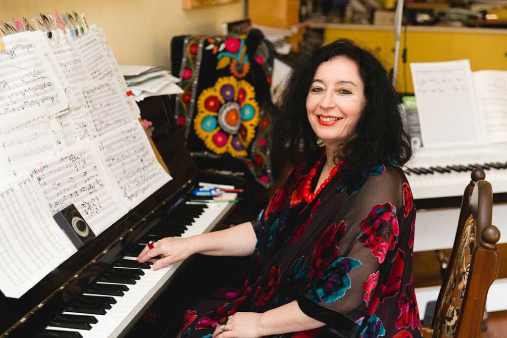 Elena-Kats-Chernin sits at a piano, smiling. Handwritten musical manuscript sits stacked on top of it.