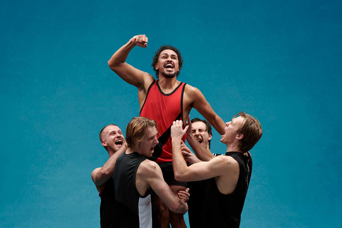 An Indigenous football player in a red jersey is lifted by four teammates in black jerseys. They all cheer.