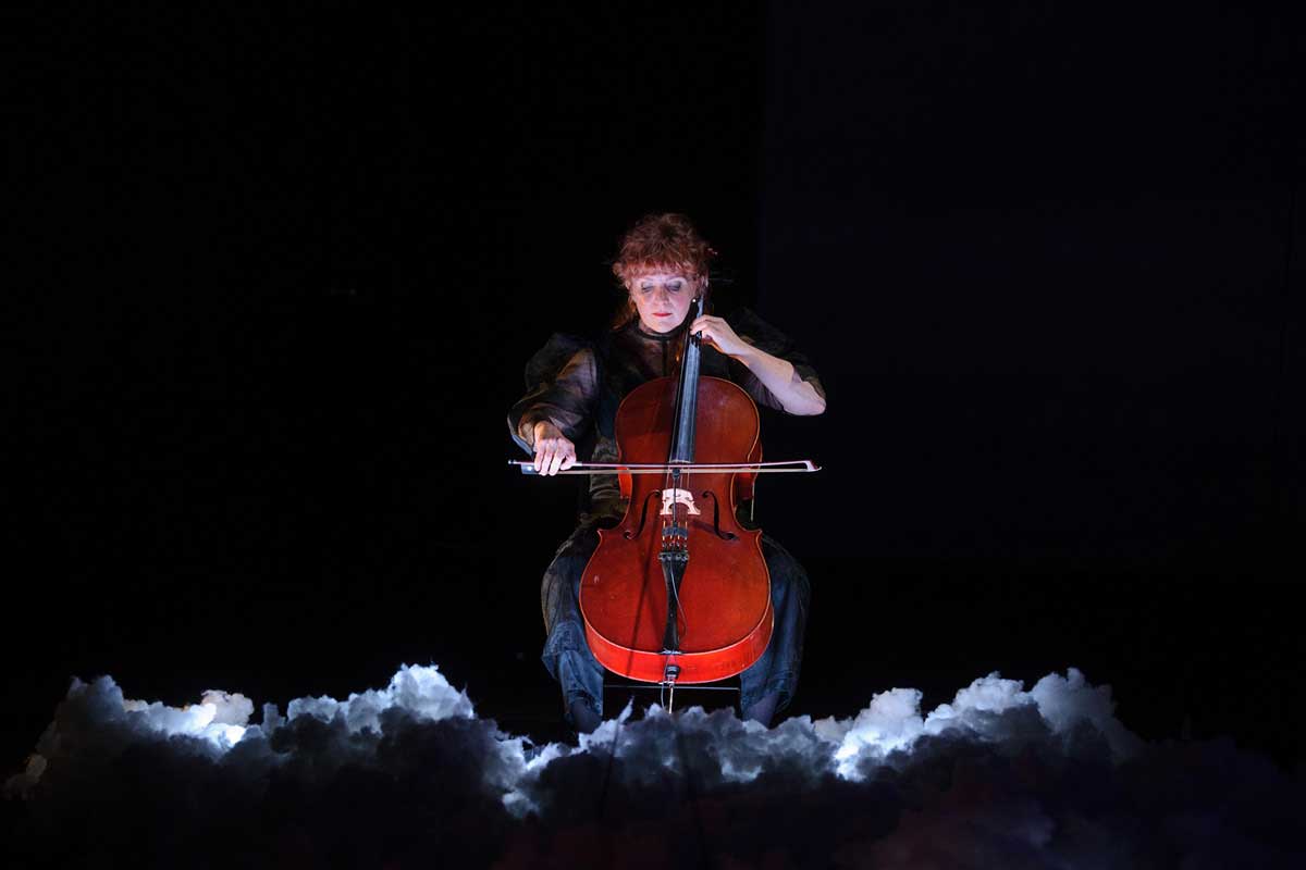 Zoe Barry plays cello above a stage floor lined with thunderclouds.