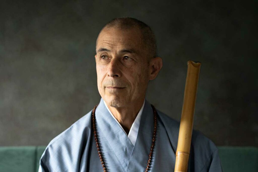 Riley Lee in a blue robe shirt, holding a Shakuhachi