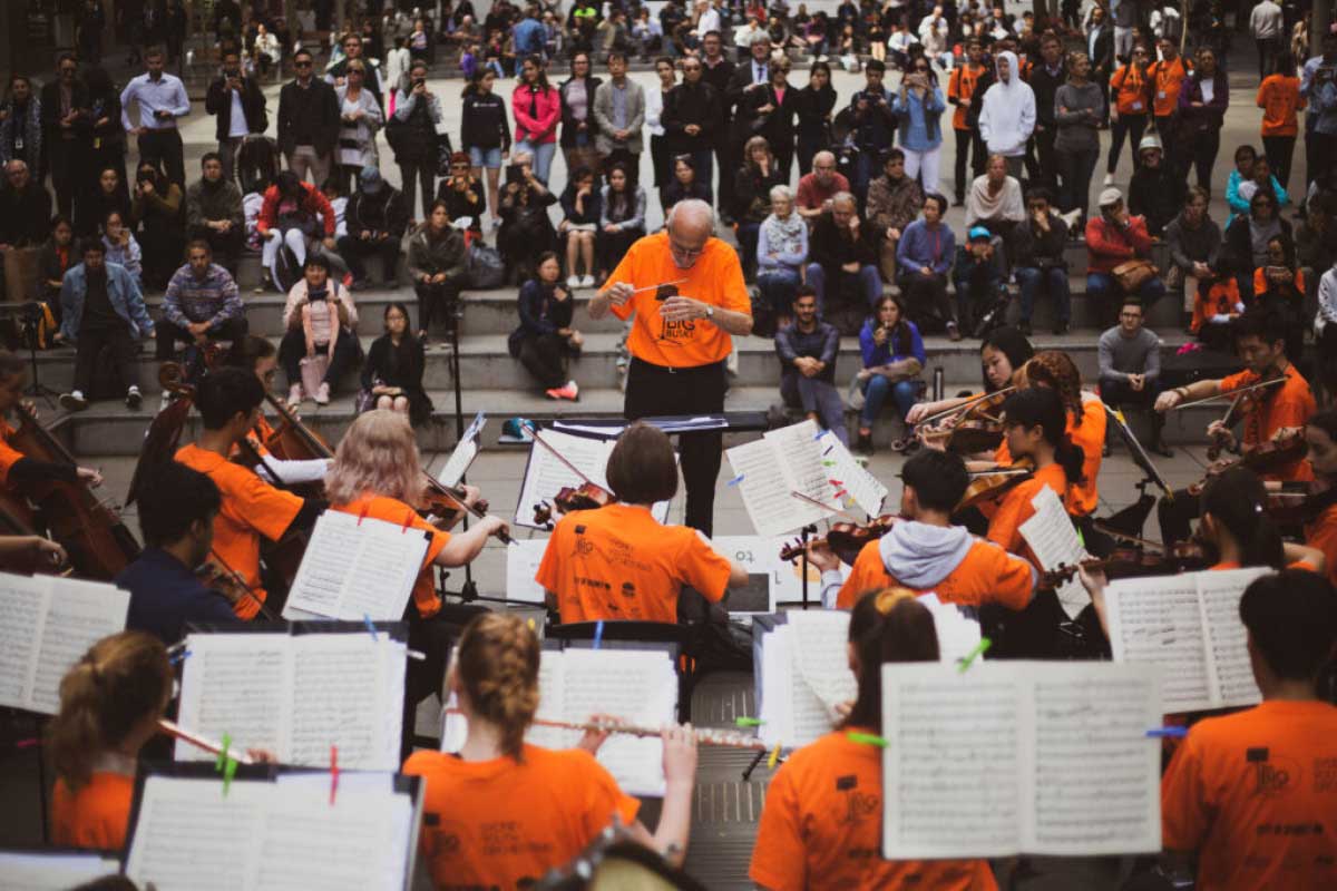 A orchestra of young musicians in bright orange shirts perform outdoors surrounded by a crowd.