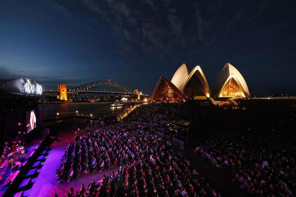 The Sydney Opera House at night, with an audience amassed on its stairs and forecourt viewing an outdoor concert.