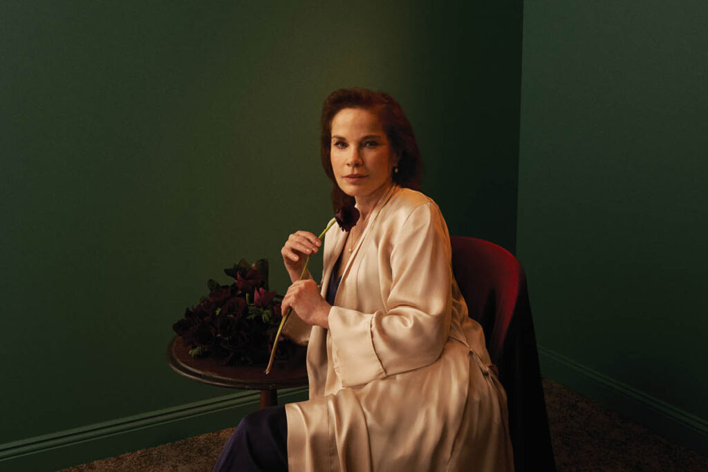 Sigrid Thornton in a white robe and slacks, holding a rose. She sits in a darkwood chair next to a table.