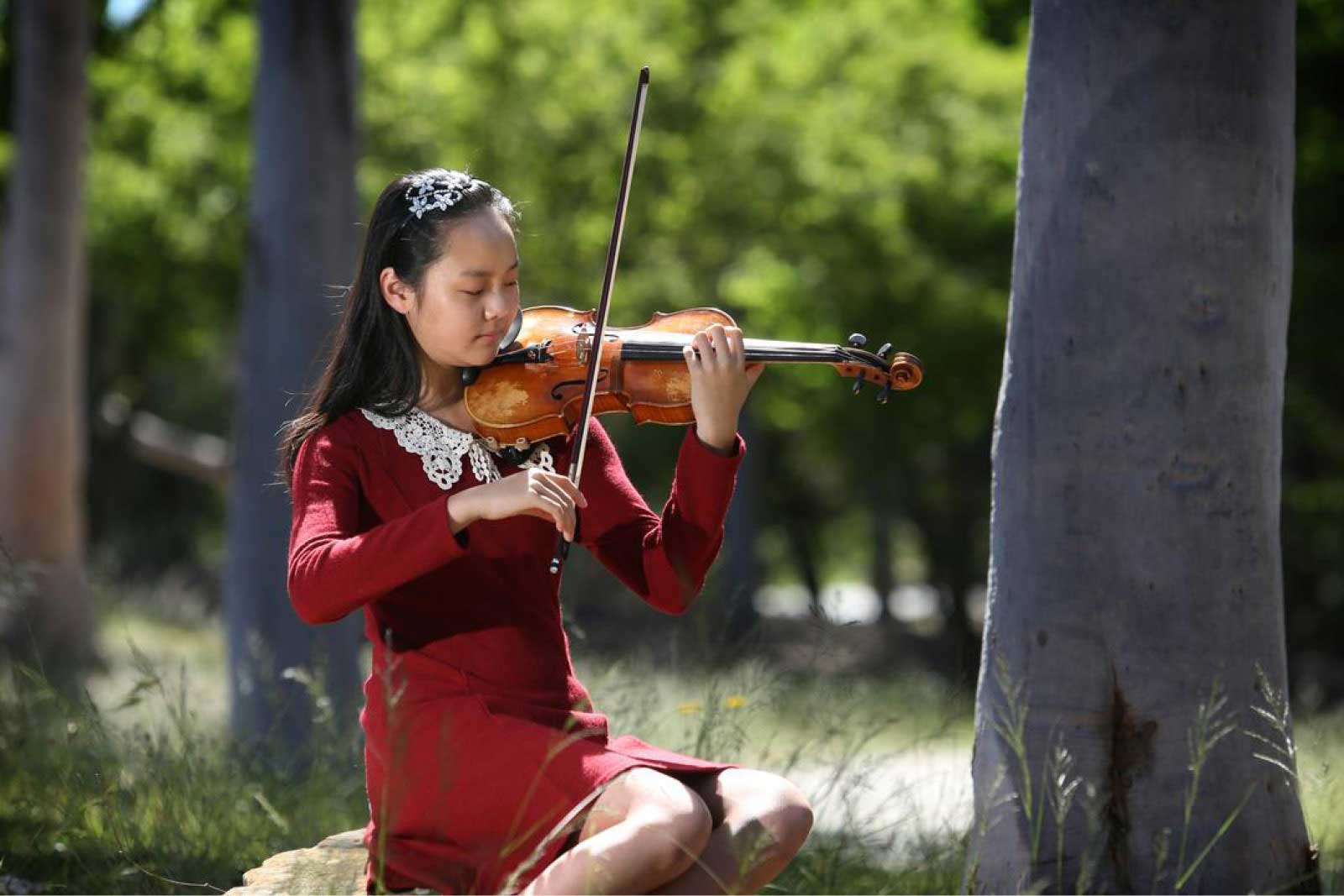 Teresa Yang sits on a rock next to a tree in a forest, playing violin.