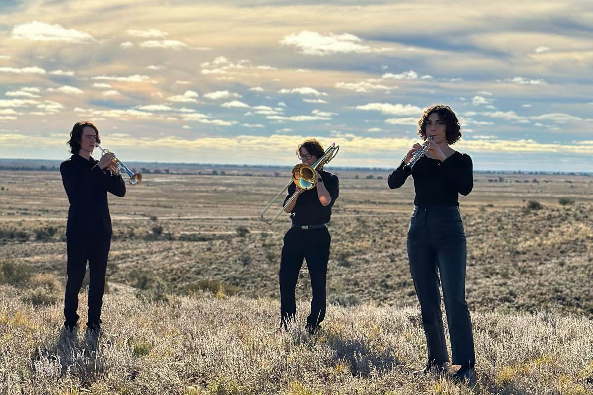 Three young musicians stand in a grassy field against a cloudy sky. One holds a trumpet, one a trombone and another an oboe.